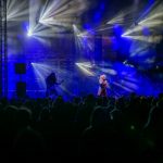 Koncert na scenie, oświetlonej niebieskimi i białymi światłami, z tłumem ludzi w ciemności. Gitarzysta po lewej, wokalista pośrodku. Dym tworzy dynamiczną atmosferę.
