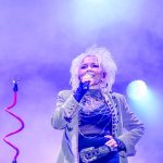 Osoba z białymi włosami śpiewa na scenie wśród fioletowych świateł. Ma na sobie ozdobną kurtkę i czarną rękawiczkę. Tło jest zamglone.
