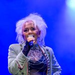 Osoba z mikrofonem na scenie, w jasnej marynarce, na tle intensywnego niebieskiego światła. Noszone duże kolczyki.