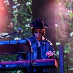 Muzyk w kapeluszu i okularach gra na keyboardzie. Tło rozmyte z motywem roślinnym, scena oświetlona kolorowymi światłami.
