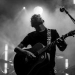 Muzyk gra na gitarze na scenie. Czarnobiałe zdjęcie pokazuje dym i światła w tle. Mikrofon widoczny z boku.