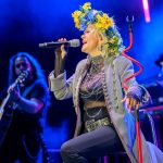 Wokalistka w wieńcu z kwiatów śpiewa na scenie. Za nią gitarzysta i klawiszowiec, scena oświetlona na niebiesko.