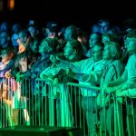 Tłum w średnim wieku ogląda występ na żywo. Kolory zieleni i błękitu oświetlają ludzi. Tło jest ciemne, co uwypukla widzów.