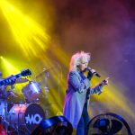 Na scenie zespół rockowy; piosenkarka w szarym płaszczu śpiewa, obok gitarzysta. Jasne, żółte światła oświetlają scenę. Tło w ciemnych barwach.