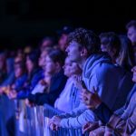 Grupa ludzi ogląda występ, skupiona na scenie. Dominują ciemne kolory z niebieskawym oświetleniem. Tło jest nieostre, podkreślając skupienie odbiorców.