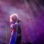 Osoba w jasnym płaszczu śpiewa na scenie w fioletowych światłach, z bębnami w tle. Dym tworzy efekt mgły. Obecne czerwone detale.