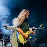 Na zdjęciu widać gitarzystę z długimi włosami na scenie, trzyma białą gitarę. Obok niego stoi osoba w jasnym stroju, z wyciągniętymi rękami. Tło jest ciemne, oświetlone niebieskim światłem i mgłą.