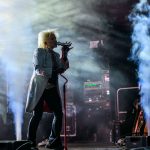 Artysta na scenie śpiewa do mikrofonu. Dym i kolorowe światła tworzą dynamiczne tło. Blond włosy i jasny strój przyciągają uwagę.