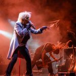 Na scenie występuje piosenkarz w jasnym płaszczu, śpiewający do mikrofonu. Obok gitarzysta z dredami, pochylony nad gitarą. Tło wypełnia czerwonawa mgła i instrumenty.