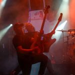 Na scenie dwóch muzyków gra na gitarach elektrycznych. Tło jest pełne intensywnego, kolorowego oświetlenia. Widać intensywnie czerwone i pomarańczowe reflektory oraz dym.