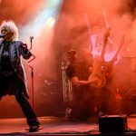 Na scenie kobieta śpiewa, trzymając mikrofon. Kolory są czerwono-pomarańczowe z efektami świetlnymi. W tle muzycy grają na gitarze i perkusji.