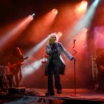 Na scenie muzyk w jasnym płaszczu śpiewa przy mikrofonie. Czerwone i białe światła oświetlają scenę. W tle dym i grający gitarzyści.