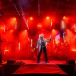 Zespół na scenie otoczony czerwonym światłem i dymem. Piosenkarka w centrum w jasnej kurtce. W tle instrumenty i efekty świetlne.