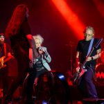 Na scenie czwórka muzyków grających rocka, w tle zestaw perkusyjny w czerwonych światłach. Gitarzysta z brodą i basista z długimi włosami. Wokalistka w szarej kurtce śpiewa do mikrofonu.