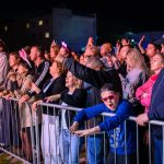 Grupa ludzi ogląda koncert za barierką nocą. Tłum jest różnorodny, widać jasno oświetlone twarze. Chłopiec w niebieskiej bluzie i czerwonych okularach wyróżnia się na pierwszym planie.