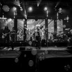 Czarno-białe zdjęcie zespołu na scenie z instrumentami, światła padają na muzyków. W tle abstrakcyjne wizualizacje. Unoszą się bańki.