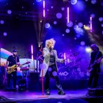 Na scenie zespół gra koncert, otoczony fioletowo-niebieskimi światłami. Wokalista śpiewa, trzymając mikrofon. Tło składa się z kolorowych świateł i efektów dymu.