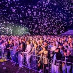 Tłum zgromadzony na koncercie w nocy z milionem mydlanych baniek. Ludzie za metalowym ogrodzeniem są podekscytowani. Światła sceniczne tworzą kolorowe iluminacje.
