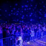 Tłum zgromadzony za barierkami na koncercie nocą. Niebieskie bąbelki unoszą się w powietrzu. Tło ciemne, zaciera szczegóły.