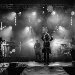 Czarno-białe zdjęcie przedstawia zespół muzyczny na scenie. W tle widać intensywne światła i dym. Muzycy grają na gitarach, perkusji i klawiszach.