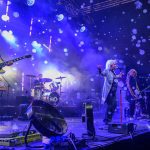 Zespół występuje na scenie przy niebieskich światłach. Muzycy grają na gitarach i perkusji, otoczeni bąbelkami. Tło jest pełne dymu i efektów świetlnych.