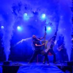Koncert rockowy z zespołem na scenie, dynamiczne światła w niebieskich tonach, efekty dymu w tle.