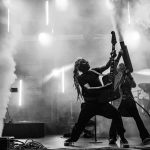 Zespół rockowy na scenie w dynamicznej pozie. Czarnobiałe zdjęcie z efektami dymu i jasnym oświetleniem w tle. Muzycy z gitarami, pełni energii.
