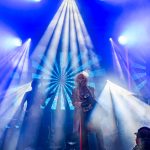 Koncert na scenie z intensywnym, niebieskim oświetleniem i promieniami światła. Wokalista śpiewa, stojąc na środku. Tło z jasnymi wzorami w kształcie wachlarza.