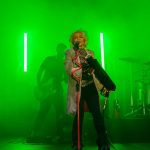 Osoba śpiewa na scenie z zielonym oświetleniem. Tło jest dymne, widoczny jest gitarzysta i perkusja. Scena ma intensywny, futurystyczny klimat.