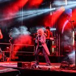 Koncert rockowy z zespołem na scenie, oświetlenie w czerwonych i niebieskich barwach. Wokalista w centrum z mikrofonem. W tle instrumenty i dym.