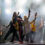 Zespół rockowy na scenie w jasnych światłach. Dwóch gitarzystów i wokalista z podniesionym mikrofonem na tle napisu Hollywood. Kolory dominujące to czerń i żółty.