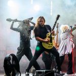 Trzech muzyków na scenie gra na gitarach. Scena oświetlona jest intensywnymi reflektorami, w tle unoszą się dymy. Centralnie mężczyzna z żółtą gitarą.