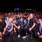 Grupa osób na scenie pozuje do zdjęcia z tłumem ludzi w tle. Jasne światła oświetlają scenę, tworząc kontrast z ciemnym niebem. Wydarzenie ma charakter koncertu lub spotkania publicznego.