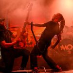 Dwóch muzyków na scenie z gitarą i perkusją, oświetlonych czerwonym światłem. W tle mocny dym. Napięta, energetyczna atmosfera.