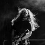 Muzyk z długimi włosami gra na gitarze elektrycznej na scenie. Czarno-białe zdjęcie z dynamicznym oświetleniem. Tło jest ciemne i zamglone.