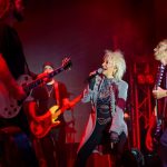 Na scenie zespół rockowy; wokalistka w jasnym płaszczu śpiewa, otoczona gitarzystami. Tło w czerwonym świetle. Klimatyczny koncert.