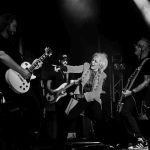 Na scenie zespół rockowy w czarno-białych barwach; gitarzysta, wokalistka i perkusista oświetleni reflektorami. Wokalistka dynamicznie gestykuluje w stronę gitarzysty. Tło zaciemnione, z widocznymi instrumentami i sprzętem koncertowym.