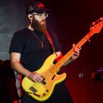 Muzyk z brodą gra na żółtej gitarze basowej na scenie. Nosi czarną czapkę i okulary. Tło jest ciemne z czerwonym oświetleniem.