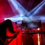 Muzyk gra na keyboardzie, w tle gitara. Dominują czerwone i niebieskie światła. Scena koncertowa pełna dymu i dramatyzmu.