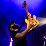 Mężczyzna gra na żółtej gitarze elektrycznej, unosząc ją w górę. Tło jest niebieskie z fioletowym oświetleniem. Scena koncertowa pełna energii.