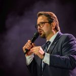 Mężczyzna w garniturze i okularach trzyma mikrofon. Tło ciemne z fioletową mgłą. Wygląda na zaangażowanego w przemowę.