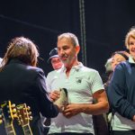 Na scenie stoi grupa osób w formalnych strojach, jedna wręcza pamiątkę. Tło czarne, z widocznymi gitarami. Kluczowe są wyrazy twarzy i interakcje.