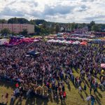 Na zdjęciu widać tłum ludzi na festynie na zielonej łące. W tle znajdują się kolorowe namioty i atrakcje, a nad nimi niebo z chmurami. Widoczne są bloki mieszkalne po lewej stronie.