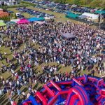 Zdjęcie przedstawia tłum ludzi na zewnątrz, uczestniczących w festiwalu. W tle scena, kolorowe namioty i zaparkowane samochody. Dominują zielone i niebieskie odcienie trawy i nieba.