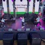 Trzech muzyków na scenie gra na instrumentach, ubrani w dżinsowe kamizelki. Scena jest oświetlona fioletowymi światłami. W tle widać duży ekran z napisami.