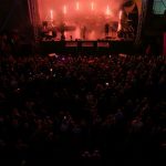 Koncert nocą: scena oświetlona czerwonym światłem, tłum ludzi na pierwszym planie. W tle ciemne niebo i namioty.