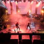 Scena koncertowa z muzykami w dymie i jasnych światłach. Przeważają kolory czerwony i pomarańczowy. Tło to ciemna przestrzeń z rampami oświetleniowymi.