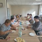 1BT6Vurn Na zdjęciu starsze kobiety siedzą przy długim drewnianym stole, jedząc pierogi. Stół jest nakryty białymi talerzami i szklanką z wodą. W tle jasne ściany i okno wpuszczające światło słoneczne.