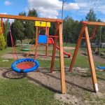 Plac zabaw z drewnianymi huśtawkami i kolorową zjeżdżalnią. Tło to trawnik, drzewa i błękitne niebo z białymi chmurami. Kolory są żywe: czerwony, niebieski i żółty.