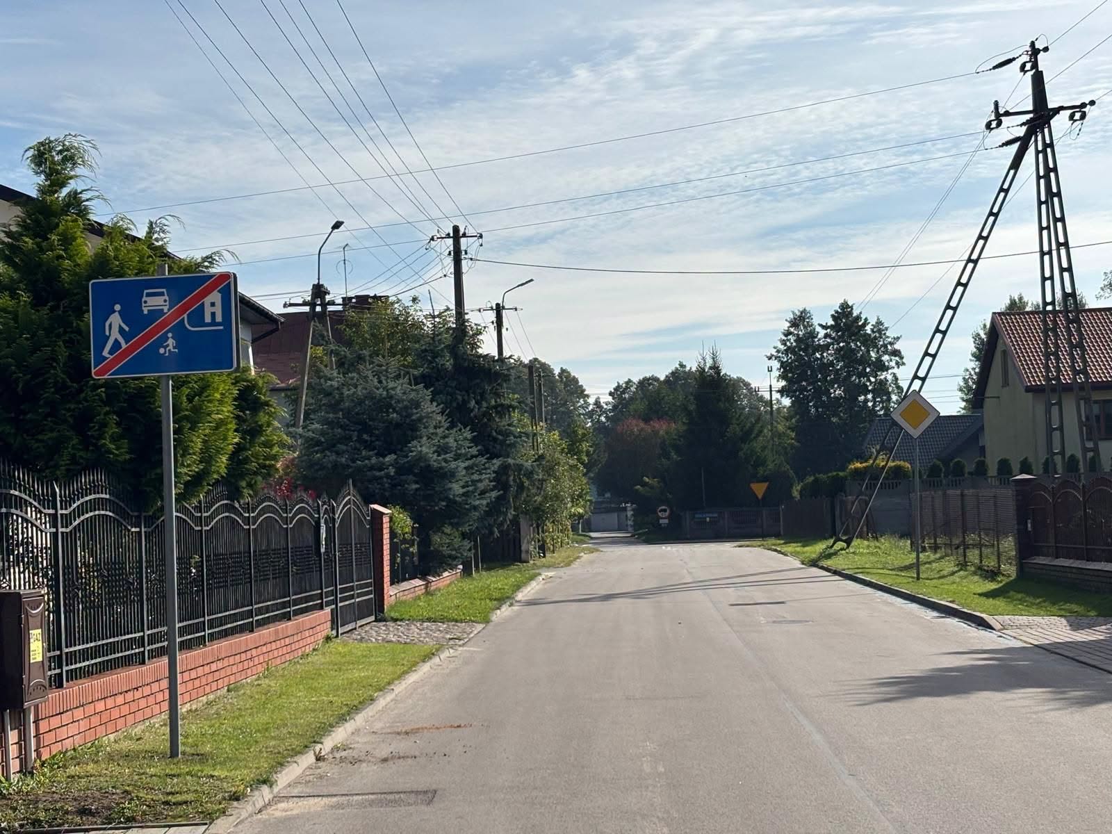 Droga w spokojnej okolicy z niebieskim znakiem końca strefy zamieszkania po lewej. Jasne domy z czerwonymi dachami w tle. Zielone drzewa i trawa wzdłuż chodnika.